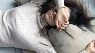 Im Bild sieht man eine liegende Frau mit einem Pulli auf einem Kissen mit einer Hand über den Augen. Vielleicht versucht sie zu schlafen.