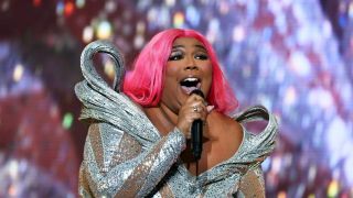 Das Bild zeigt Lizzo mit pinken Haaren und einem silbernen Kleid auf der Bühne beim Singen.