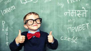 Ein junger Bub mit zwei erhobenen Daumen lächelt mit geschlossenen Augen und einer viel zu großen Brillei n die Kamera. Hinter ihm ist eine grüne Tafel mit verschiedenen Wörtern in verschiedenen Sprachen: lorem, dia duit, ahoj, hoy, etc.