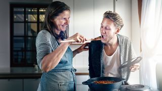Ein ungefähr 25-jähriger Mann mit offenem Mund wird von seiner Mutter von einem Kochlöffel gefüttert. Die Beiden stehen an einem Herd, wo ein Topf mit Soße steht. Die Beiden lächeln.