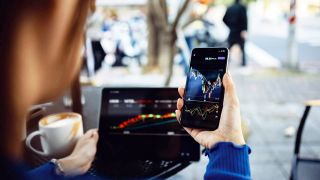 Im Bild sieht man die Hand einer Person, die ein Handy in der Hand hält und in einem Cafe sitzt. Auf dem Handy und am Laptop sind Aktienkursentwicklungen zu sehen. Die ganze Szene ist in einem Cafe im Freien mit Menschen im Hintergrund.