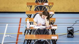 Im Bild siehst du ein Mädchen das einen Prüfungsbogen in einer Turnhalle ausfüllt. Sie sitzt auf einem einzelnen Tisch, hinter ihr sieht man zwei leere Tische und vor ihr einen leeren Tisch.