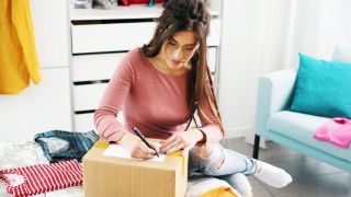 Eine junge Frau mit langen dunklen Haaren und Brille sitzt auf der Couch und beschriftet ein Paket.