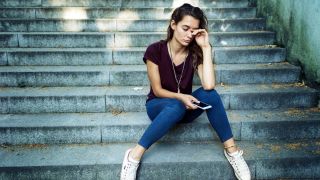 Im Bild sitzt eine Frau mit einem Handy in der Hand auf einer Treppe. Sie sieht nachdenklich und traurig bis verzweifelt aus.